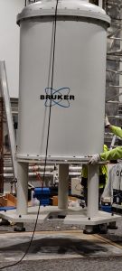 A large white Bruker 750 MHz NMR magnet stands on its support legs while personnel removes it from place. Cables, tools, and rigging equipment are visible around the instrument.
