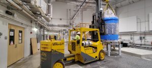 A yellow Versa-Lift forklift inside a laboratory hall raises the crated 1.1 GHz NMR magnet, wrapped in blue protective material, while rigging straps support it from overhead. The scene shows the lift maneuvering the magnet near a concrete instrument pad with facility piping and equipment in the background.