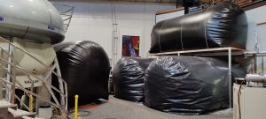 Large black gas‑collection bags for the liquid helium recovery system are arranged on the floor and on an elevated platform next to a tall white 900 MHz NMR magnet inside a lab instrumentation room.