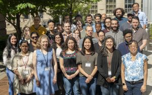 A large group of workshop attendees stands outdoors in front of a building and trees during the 2024 Solution NMR Workshop. The group is arranged in several rows, many wearing name badges, and positioned together for an event photo.
