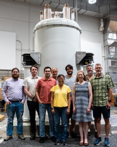 A group of NMRFAM personnel stands in front of a large, white 1.1 GHz NMR magnet inside the facility. The magnet has structural components and wiring visible near the top section, and the group is positioned directly in front of the instrument for a documentation photo.