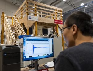 A person sits at a workstation reviewing NMR data displayed on a computer monitor, which shows a blue spectral trace. In the background, the wooden support platform for the 1.1 GHz NMR magnet is visible, with stairs and signage mounted on the structure.