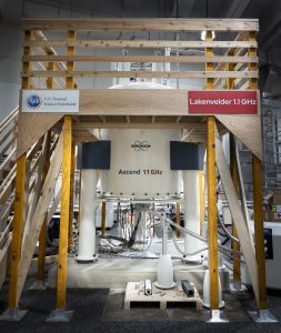 Front view of a large Bruker Ascend 1.1 GHz NMR magnet housed within a tall wooden support platform. The structure includes stairs on both sides and signage from the U.S. National Science Foundation and “Lakenvelder 1.1 GHz.” Cables and instrumentation are visible beneath the magnet on the floor.