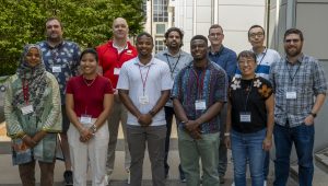 A group photo of workshop attendees standing outdoors in front of a building and greenery. The group includes multiple individuals wearing name badges, arranged in two rows for the 2024 SSNMR Workshop.